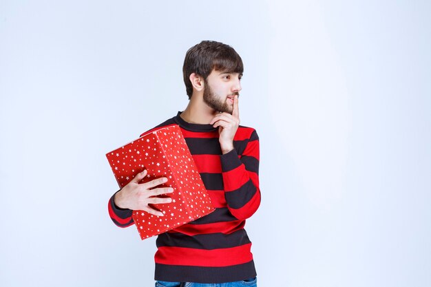 Homem de camisa listrada vermelha segurando uma caixa de presente vermelha e parece confuso e pensativo.