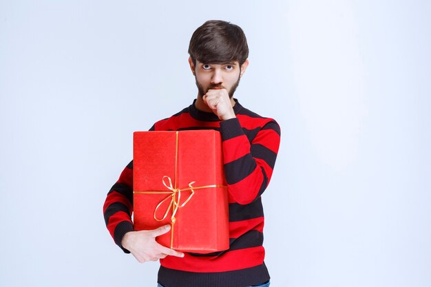 Homem de camisa listrada vermelha segurando uma caixa de presente vermelha e parece confuso e pensativo.