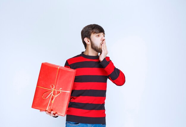 Homem de camisa listrada vermelha, segurando uma caixa de presente vermelha, parece cansado e com sono.