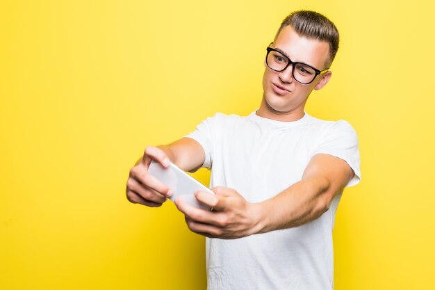 Homem de camiseta branca e óculos faz algo em seu telefone e tira fotos de selfie isoladas em amarelo