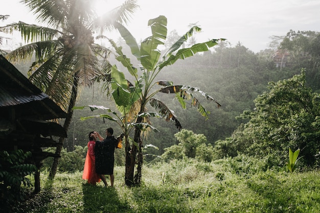 Homem de capa de chuva preta tocando o rosto da namorada na natureza. Dois turistas posando na floresta tropical.