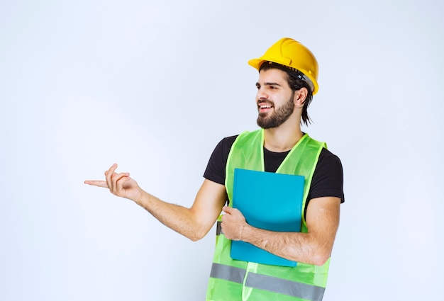 Homem de capacete segurando uma pasta azul e apontando alguém à esquerda.