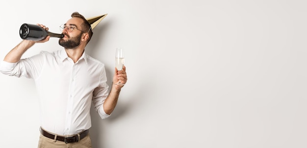 Homem de celebração e feriados bebendo chamapgne de garrafa na festa de aniversário em pé contra branco