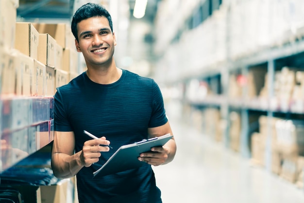 Homem de engenheiro indiano inteligente usando capacete de segurança fazendo verificação de estoque e gerenciamento de produtos de estoque de papelão no fundo do armazém da fábrica