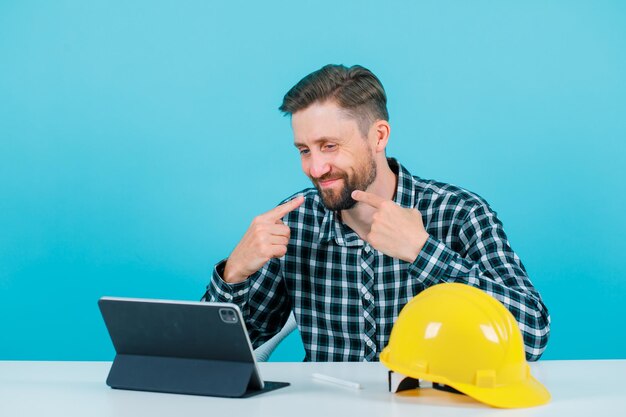 Homem de engenheiro sorridente está mostrando seu sorriso com os dedos, sentado na frente do tablet em fundo azul