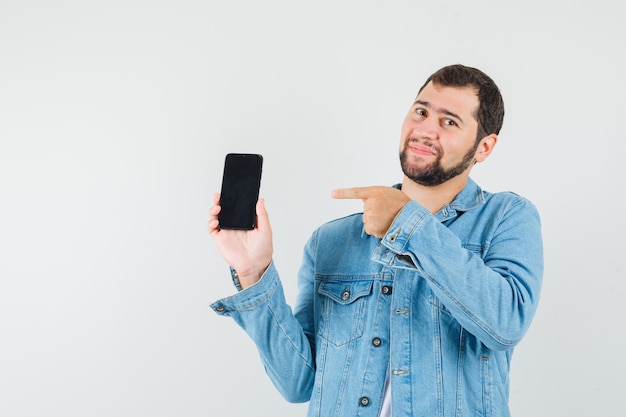 Foto grátis homem de estilo retro apontando para o celular com jaqueta, camiseta e parecendo satisfeito. vista frontal.