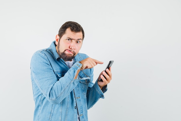 Foto grátis homem de estilo retro com casaco, t-shirt marcante com a língua de fora enquanto toca a tela do telefone e olhando concentrado, vista frontal