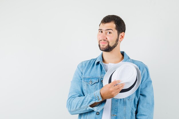 Homem de estilo retro com jaqueta, camiseta segurando o chapéu branco e parecendo calmo, vista frontal.