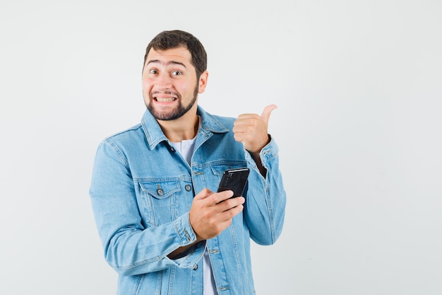 Homem de estilo retro com jaqueta, t-shirt apontando para longe, segurando o telefone e olhando alegre, vista frontal.