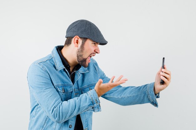 Homem de estilo retro falando alguma coisa enquanto faz videochamada com jaqueta, boné, camisa e parece agressivo.