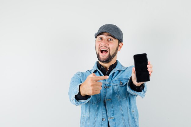 Homem de estilo retro na jaqueta, boné, camisa, apontando para o telefone e olhando alegre, vista frontal.