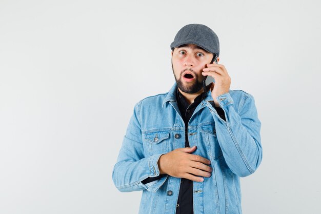 Homem de estilo retro na jaqueta, boné, camisa, falando no celular e olhando com medo, vista frontal. espaço para texto