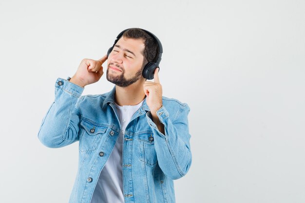 Homem de estilo retro ouvindo música com fones de ouvido na jaqueta, camiseta e parecendo relaxado, vista frontal.