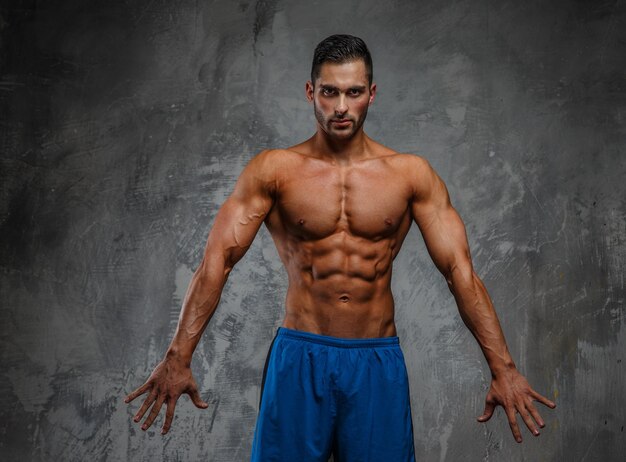 Homem de fitness sem camisa muscular em shorts azuis posando no estúdio em fundo cinza