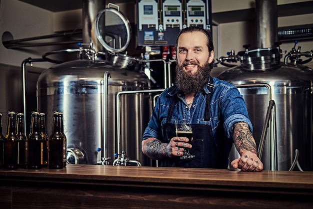 Homem de hipster tatuado e barbudo sorridente em uma camisa jeans e avental trabalhando em uma fábrica de cervejaria, atrás de um balcão, segura um copo de cerveja para controle de qualidade.