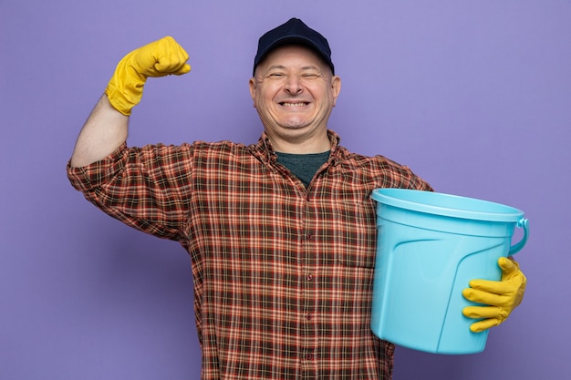 Homem de limpeza com camisa xadrez e boné, usando luvas de borracha, segurando um balde, levantando o punho, feliz e animado como um vencedor em pé sobre um fundo roxo