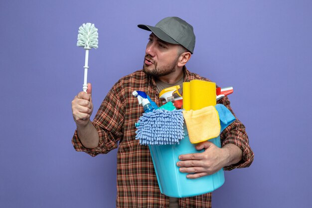 Homem de limpeza eslavo confuso segurando equipamento de limpeza e olhando para a escova de banheiro