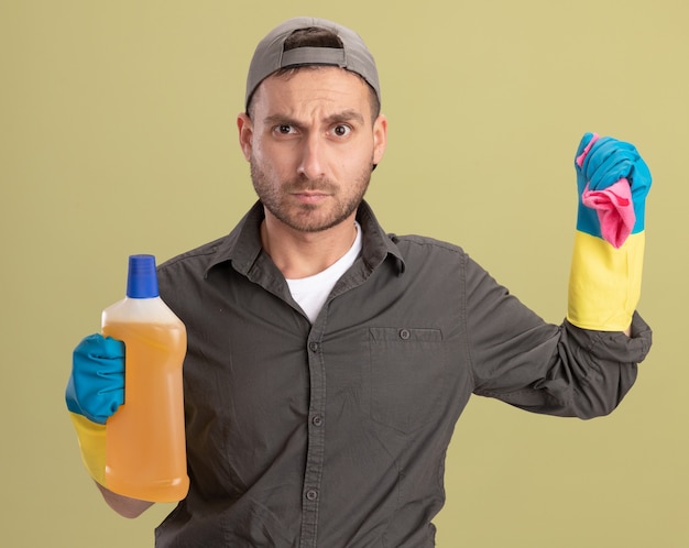 Homem de limpeza jovem vestindo roupas casuais e boné em luvas de borracha segurando uma garrafa com material de limpeza e um pano olhando com cara de raiva em pé sobre a parede verde