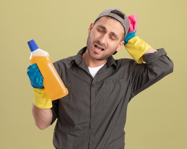 Homem de limpeza jovem vestindo roupas casuais e boné em luvas de borracha segurando uma garrafa com material de limpeza e um pano, parecendo confuso e muito ansioso em pé sobre a parede verde