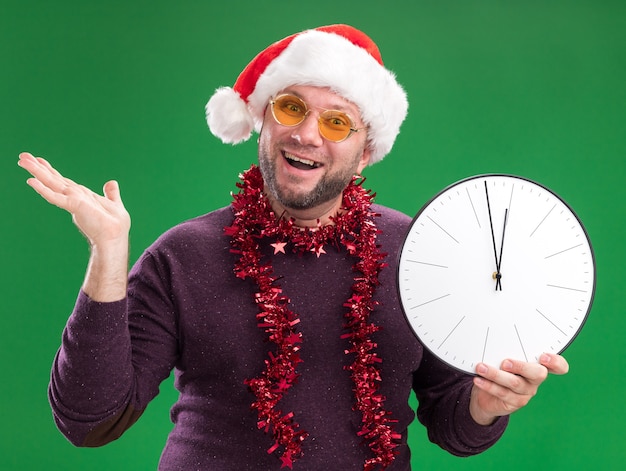 Homem de meia-idade alegre com chapéu de Papai Noel e guirlanda de ouropel no pescoço, óculos segurando um relógio e mostrando a mão vazia isolada na parede verde