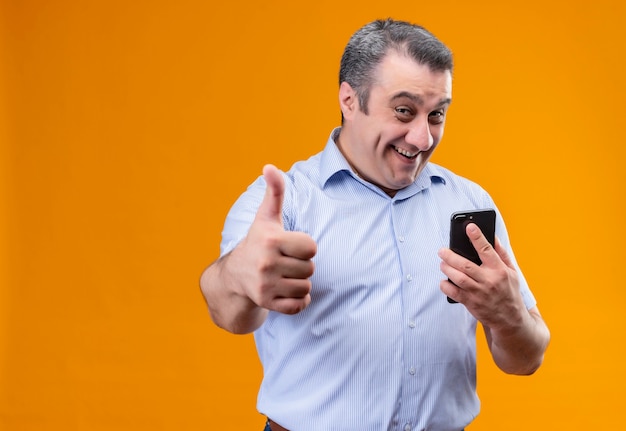 Homem de meia-idade alegre e positivo, vestindo uma camisa azul listrada, segurando um telefone celular e mostrando os polegares para cima