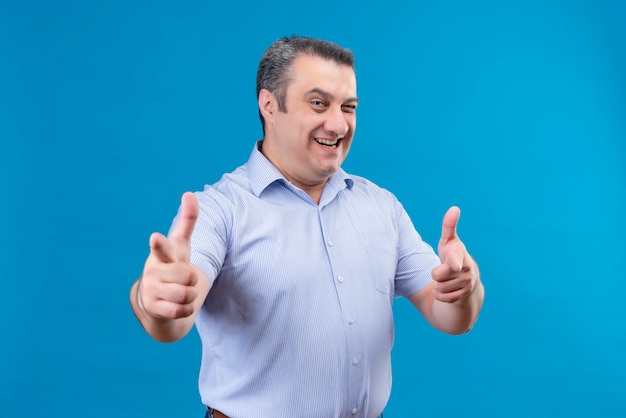 Homem de meia-idade alegre e sorridente com camisa listrada azul, apontando com o dedo indicador e piscando para a câmera sobre um fundo azul