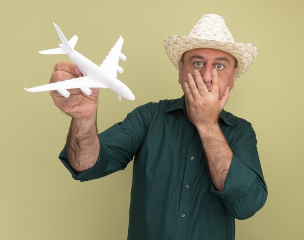 Homem de meia-idade assustado vestindo camiseta e chapéu verdes segurando um avião de brinquedo e colocando a mão na boca isolada na parede verde oliva