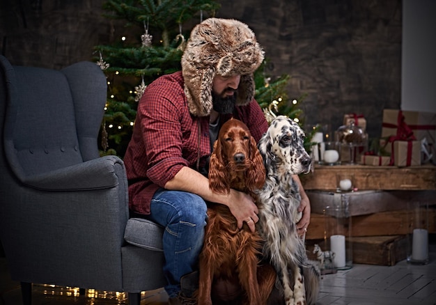 Homem de meia idade barbudo vestido com uma camisa xadrez e chapéu de pele e dois cães setter irlandês com árvore de abeto e decoração de Natal no fundo.