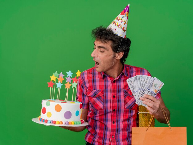 Homem de meia idade, caucasiano, festeiro, usando um boné de aniversário, segurando um bolo de aniversário, sacola de papel, pacote de presente e dinheiro olhando para o bolo se preparando para morder