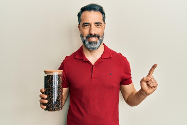 Homem de meia-idade com barba e cabelos grisalhos segurando jarro com grãos de café sorrindo feliz apontando com a mão e o dedo para o lado