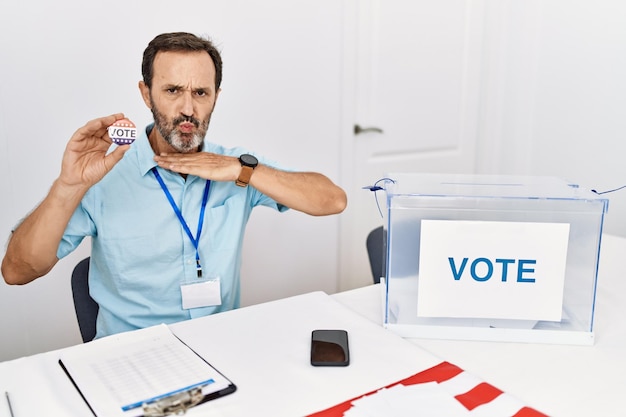 Homem de meia-idade com barba sentado perto da cédula segurando o distintivo de voto cortando a garganta com a mão enquanto faca ameaça agressão com violência furiosa