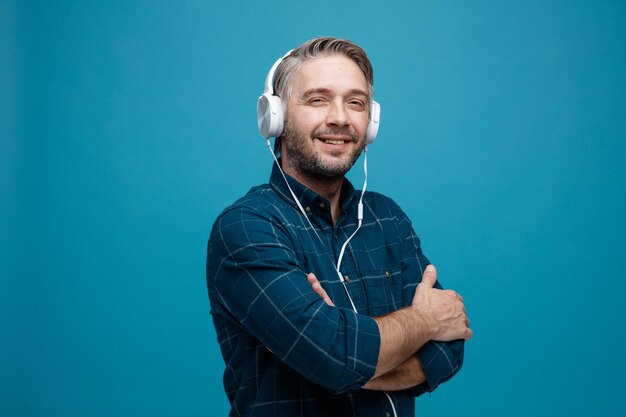 Homem de meia idade com cabelos grisalhos na camisa de cor escura com fones de ouvido, olhando para a câmera com um grande sorriso no rosto, cruzando os braços no peito em pé sobre fundo azul