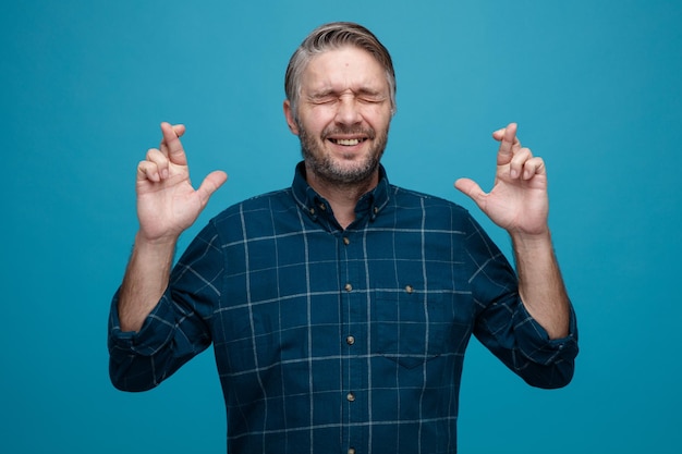 Foto grátis homem de meia-idade com cabelos grisalhos na camisa de cor escura, cruzando os dedos, desejando ser animado em pé sobre fundo azul