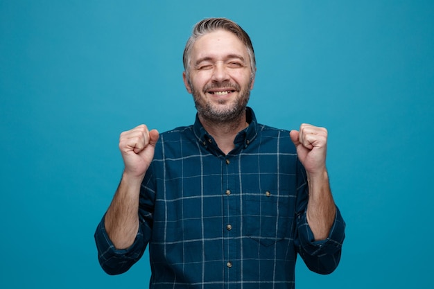 Foto grátis homem de meia idade com cabelos grisalhos na camisa de cor escura feliz e animado, cerrando os punhos, regozijando-se com seu sucesso em pé sobre fundo azul