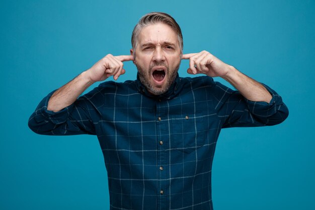 Homem de meia idade com cabelos grisalhos na camisa de cor escura gritando fechando os ouvidos com os dedos com expressão irritada em pé sobre fundo azul