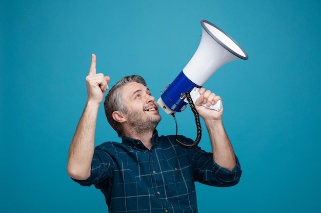 Foto grátis homem de meia idade com cabelos grisalhos na camisa de cor escura gritando no megafone sendo animado e feliz olhando para cima apontando com o dedo indicador em pé sobre fundo azul