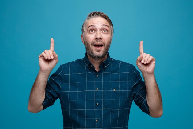 homem de meia idade com cabelos grisalhos na camisa de cor escura olhando feliz e surpreso apontando com os dedos indicadores em pé sobre fundo azul