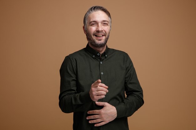 Homem de meia idade com cabelos grisalhos na camisa de cor escura olhando para algo rindo apontando com o dedo indicador para algo ou alguém de pé sobre fundo marrom