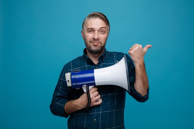 Homem de meia idade com cabelos grisalhos na camisa de cor escura segurando o megafone olhando para a câmera sorrindo feliz e positivo apontando com o polegar para o lado em pé sobre fundo azul