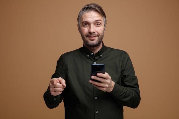 homem de meia idade com cabelos grisalhos na camisa de cor escura, segurando o smartphone apontando com o dedo indicador para a câmera sorrindo confiante, feliz e positivo em pé sobre fundo marrom