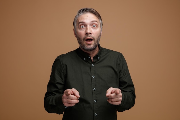 Homem de meia idade com cabelos grisalhos na camisa verde escura, olhando para a câmera espantado e surpreso apontando com os dedos indicadores para a câmera em pé sobre fundo marrom