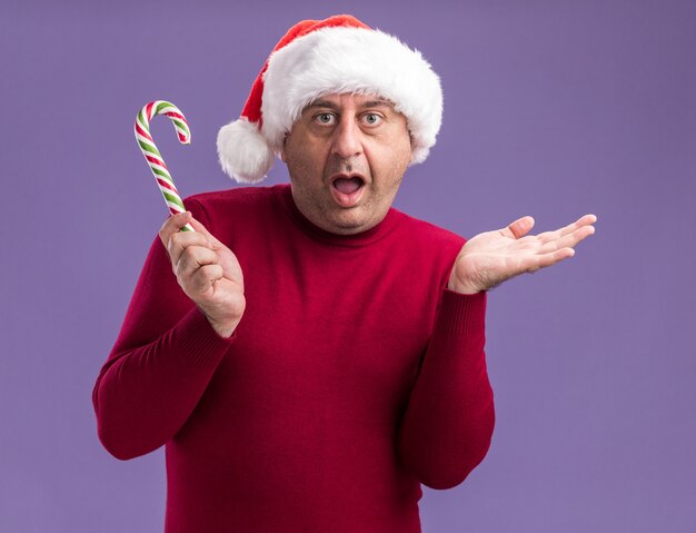 Homem de meia-idade com chapéu de Papai Noel de Natal segurando uma bengala de doces feliz e surpreso com o braço levantado em pé sobre a parede roxa