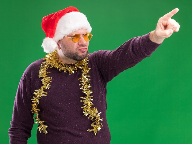 Homem de meia-idade confiante usando chapéu de Papai Noel e guirlanda de ouropel no pescoço, óculos, olhando e apontando para o lado isolado no fundo verde