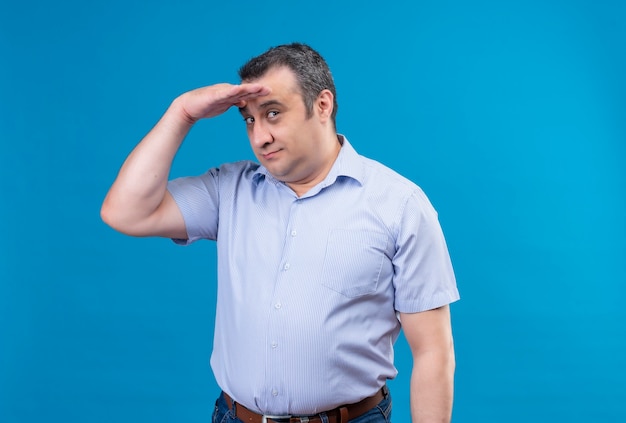 Homem de meia-idade confuso com camisa listrada azul, olhando para longe, segurando a testa em um espaço azul