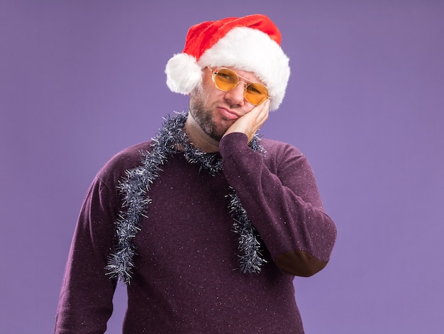 Homem de meia-idade confuso com chapéu de Papai Noel e guirlanda de ouropel no pescoço, óculos mantendo a mão no rosto isolado na parede roxa