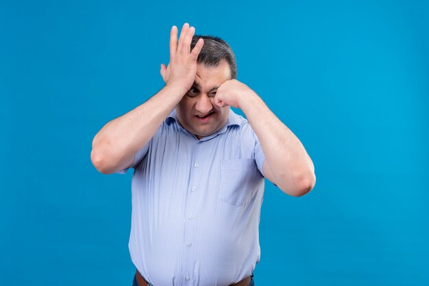 Homem de meia-idade confuso e chateado com uma camisa listrada vertical azul, segurando a mão na testa em um espaço azul