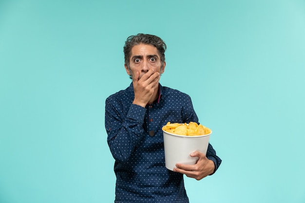 Homem de meia-idade, de frente, segurando uma cesta com cips e assistindo a um filme na superfície azul