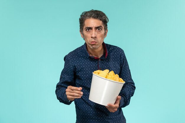 Homem de meia-idade de vista frontal com uma cesta cheia de cips e comendo em uma superfície azul clara