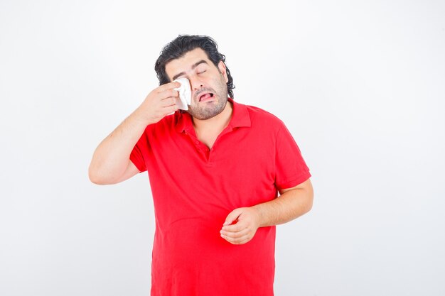 Homem de meia idade em t-shirt vermelha, enxugando os olhos com o guardanapo enquanto chora e parece ofendido, vista frontal.