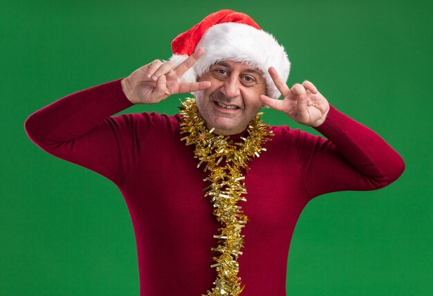 Homem de meia-idade feliz com chapéu de Papai Noel de Natal com enfeites no pescoço e sorrindo mostrando o sinal V perto dos olhos em pé sobre a parede verde